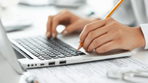 Handen typen op laptop met potlood in rechterhand; papieren eronder, kantoor, prompt engineering tips voor professionals.