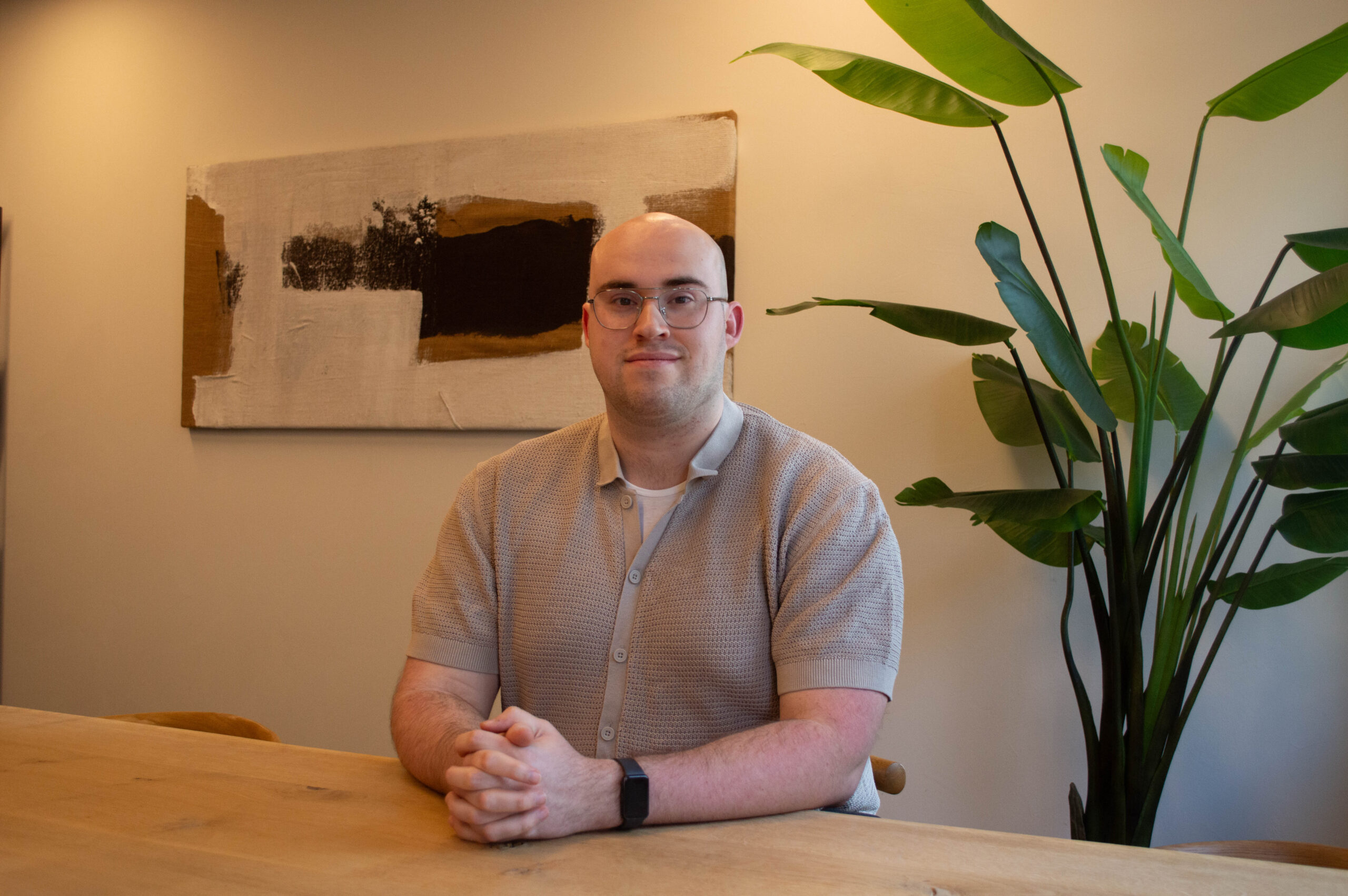 Man met bril zit aan een houten tafel in een rustige ruimte met abstract schilderij en grote groene plant.