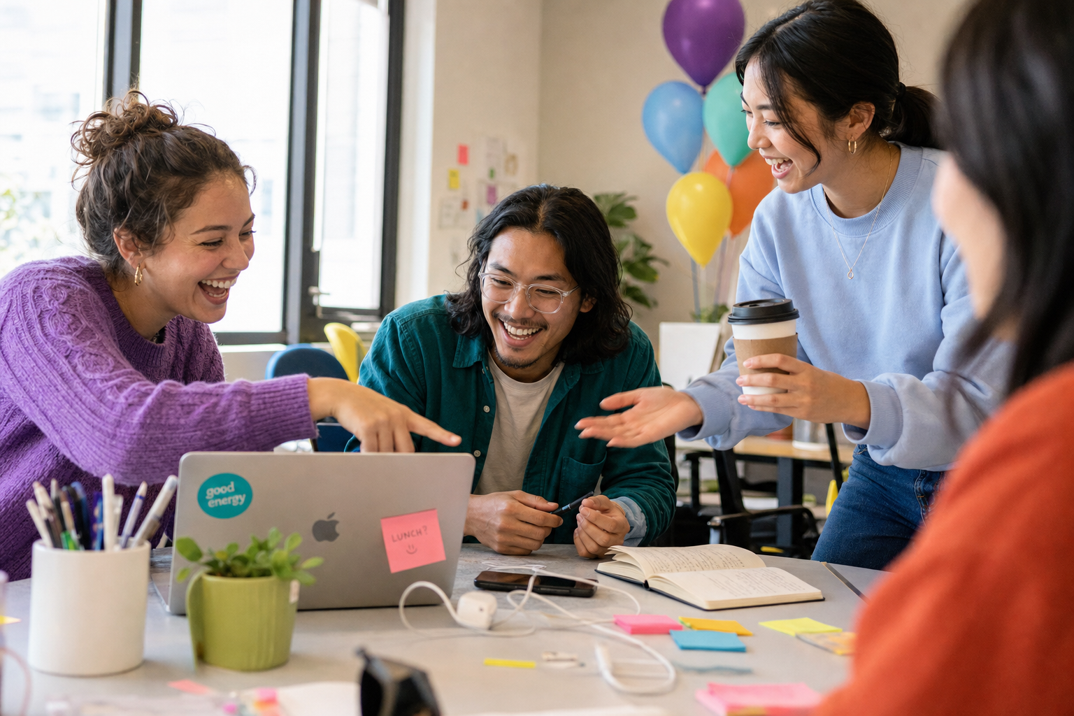 Drie collega's lachen en overleggen rond een laptop aan een bureau met notitieboek, post-its en ballonnen.