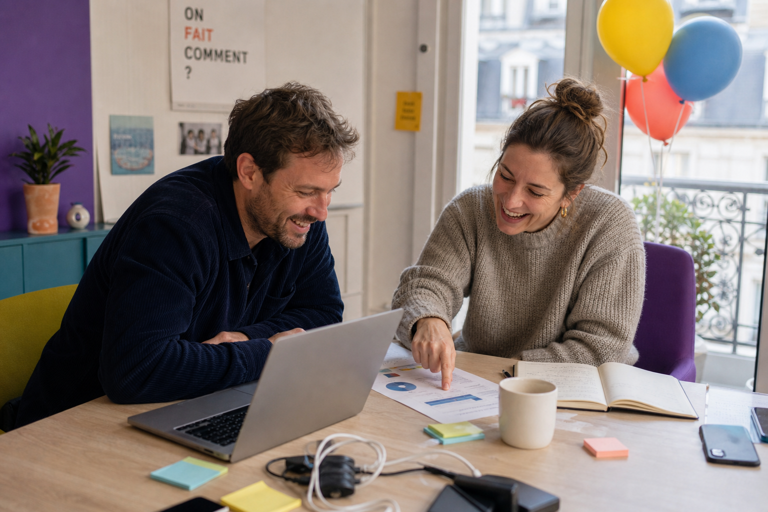 Twee collega's lachen aan tafel terwijl ze samen een grafiek en laptop bekijken op kantoor.