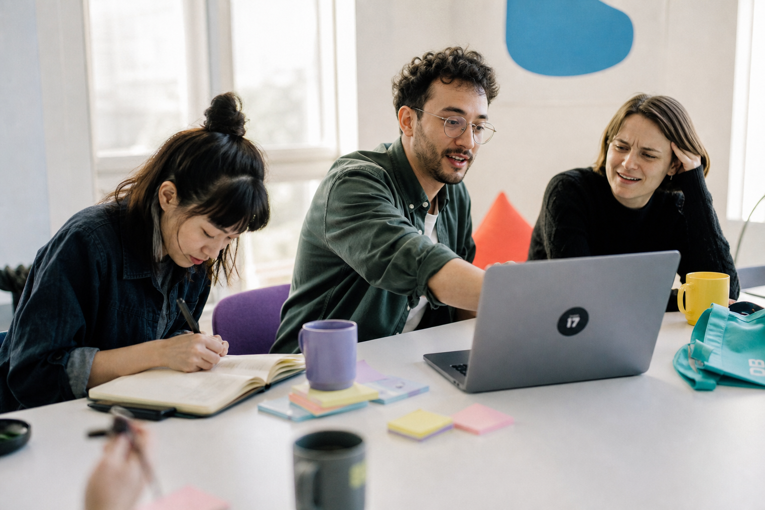 Drie collega’s overleggen aan een tafel met laptop, notitieboek en gekleurde mokken in een lichte kantoorruimte.