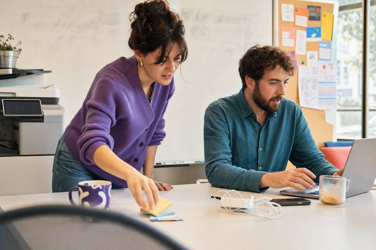 Twee collega’s werken samen aan een laptop in een kantoorruimte met whiteboard en notities op de achtergrond.
