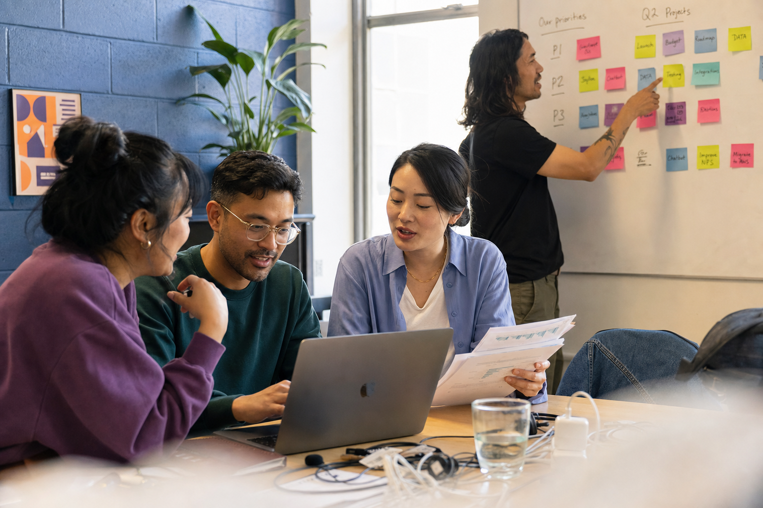Vier collega's overleggen aan tafel met een laptop, terwijl een collega sticky notes op een whiteboard aanwijst.